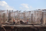 Workers are on a construction site with metal scaffolding rising against a blue sky. The workers are wearing safety gear including helmets and vests, and they appear to be engaged in various tasks. The setting suggests an active building project with visible infrastructure and stacks of construction materials around.