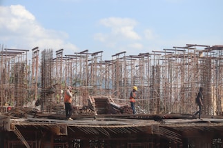 Workers are on a construction site with metal scaffolding rising against a blue sky. The workers are wearing safety gear including helmets and vests, and they appear to be engaged in various tasks. The setting suggests an active building project with visible infrastructure and stacks of construction materials around.