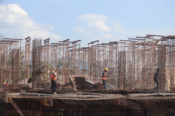 Workers are on a construction site with metal scaffolding rising against a blue sky. The workers are wearing safety gear including helmets and vests, and they appear to be engaged in various tasks. The setting suggests an active building project with visible infrastructure and stacks of construction materials around.