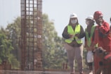 A group of construction workers wearing safety gear, including helmets and reflective vests, are gathered near a vertical metal structure on a construction site. Trees and greenery are visible in the background.
