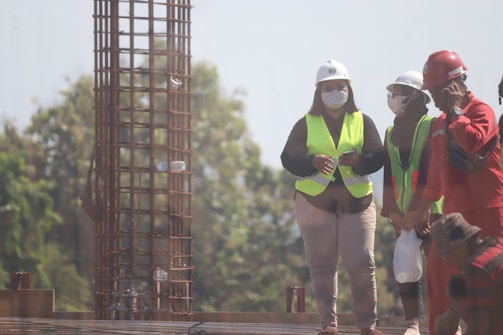 A group of construction workers wearing safety gear, including helmets and reflective vests, are gathered near a vertical metal structure on a construction site. Trees and greenery are visible in the background.