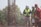 A group of construction workers wearing safety gear, including helmets and reflective vests, are gathered near a vertical metal structure on a construction site. Trees and greenery are visible in the background.