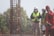 A group of construction workers wearing safety gear, including helmets and reflective vests, are gathered near a vertical metal structure on a construction site. Trees and greenery are visible in the background.