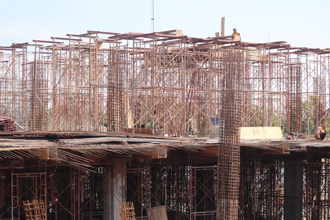 Scaffolding and construction materials form a complex structure, with metallic frameworks rising vertically. Several workers are present, engaging in construction tasks on an unfinished building.