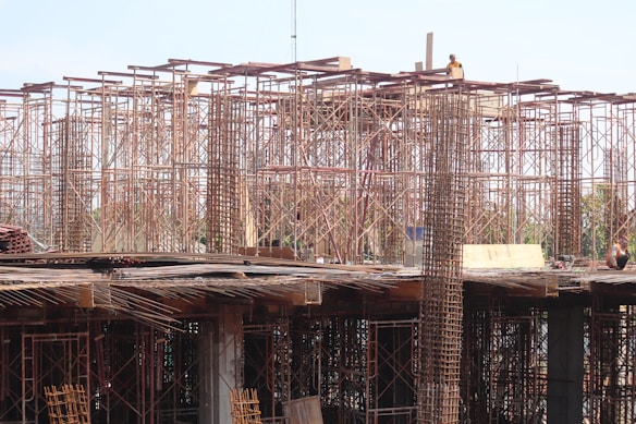Scaffolding and construction materials form a complex structure, with metallic frameworks rising vertically. Several workers are present, engaging in construction tasks on an unfinished building.