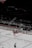 An ice hockey match in a stadium with players in red and white uniforms on the ice rink. Advertisements and boards line the rink's perimeter. A large blimp with Cyrillic writing hovers above the seating area.