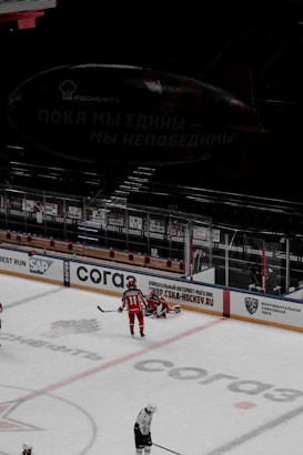 An ice hockey match in a stadium with players in red and white uniforms on the ice rink. Advertisements and boards line the rink's perimeter. A large blimp with Cyrillic writing hovers above the seating area.