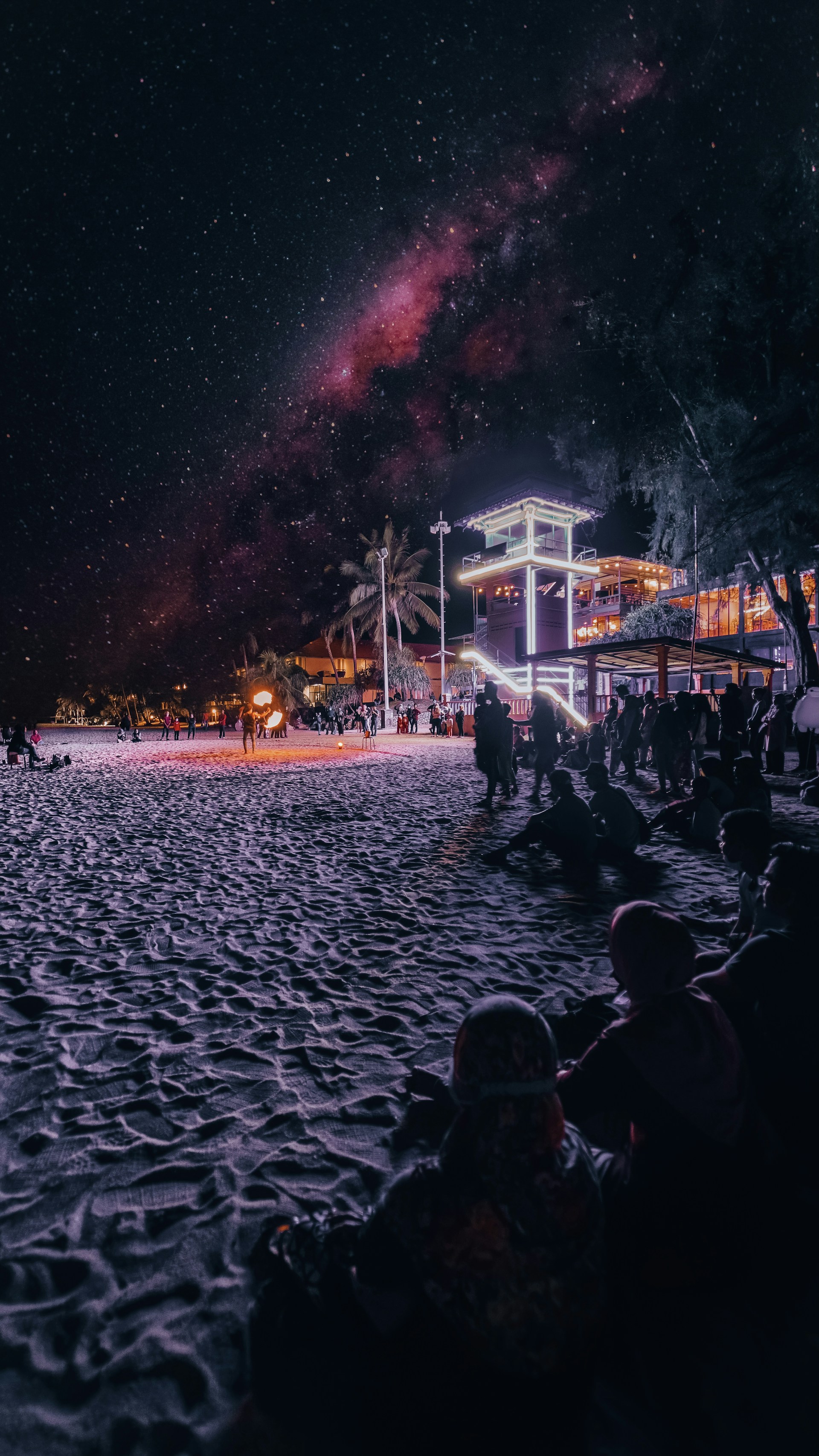 A nighttime beach scene with people gathered, possibly watching a performance or enjoying the atmosphere. The sky is full of stars and the Milky Way is visible, creating a surreal backdrop. A brightly lit structure with neon lights is present near the shoreline, adding a modern touch to the natural setting.