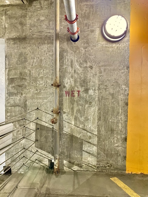 A dimly lit concrete wall with a large metal pipe running vertically next to it. A round light fixture is mounted on the wall casting shadows. The word 'WET' is stenciled in red on the wall. A railing runs along the stairway on the left.