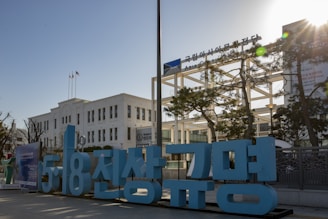 A white building with multiple windows, likely an official or historical structure, stands prominently. In the foreground, there are large blue letters in a different language. Two flags are visible atop a pole, and a sign labeled 'Asia Culture Center' is seen. Several trees and a clear blue sky fill the background.