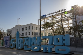 A white building with multiple windows, likely an official or historical structure, stands prominently. In the foreground, there are large blue letters in a different language. Two flags are visible atop a pole, and a sign labeled 'Asia Culture Center' is seen. Several trees and a clear blue sky fill the background.
