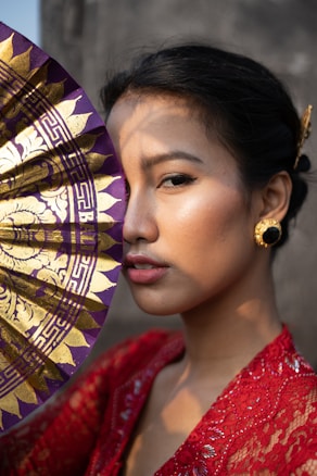 A person with elegant makeup is holding a decorative fan that partially covers the face. They are wearing ornate gold earrings and a bright red, textured garment. The lighting casts a warm golden hue, highlighting the person's features and adding depth to the image.