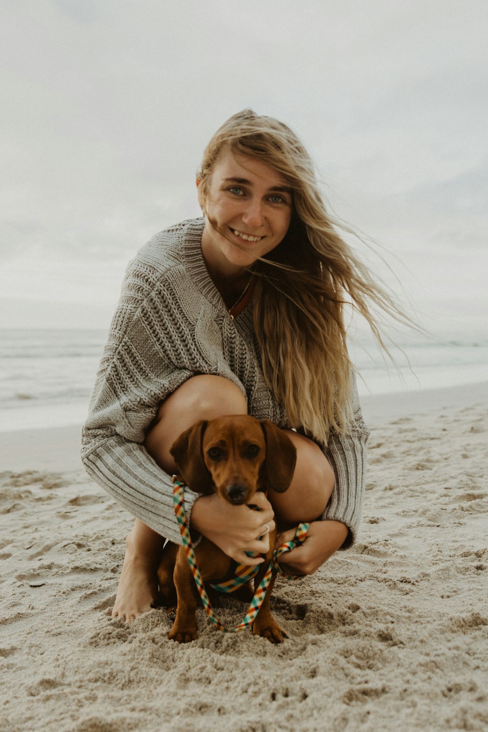 woman in gray knit sweater hugging brown short coated dog on beach during daytime