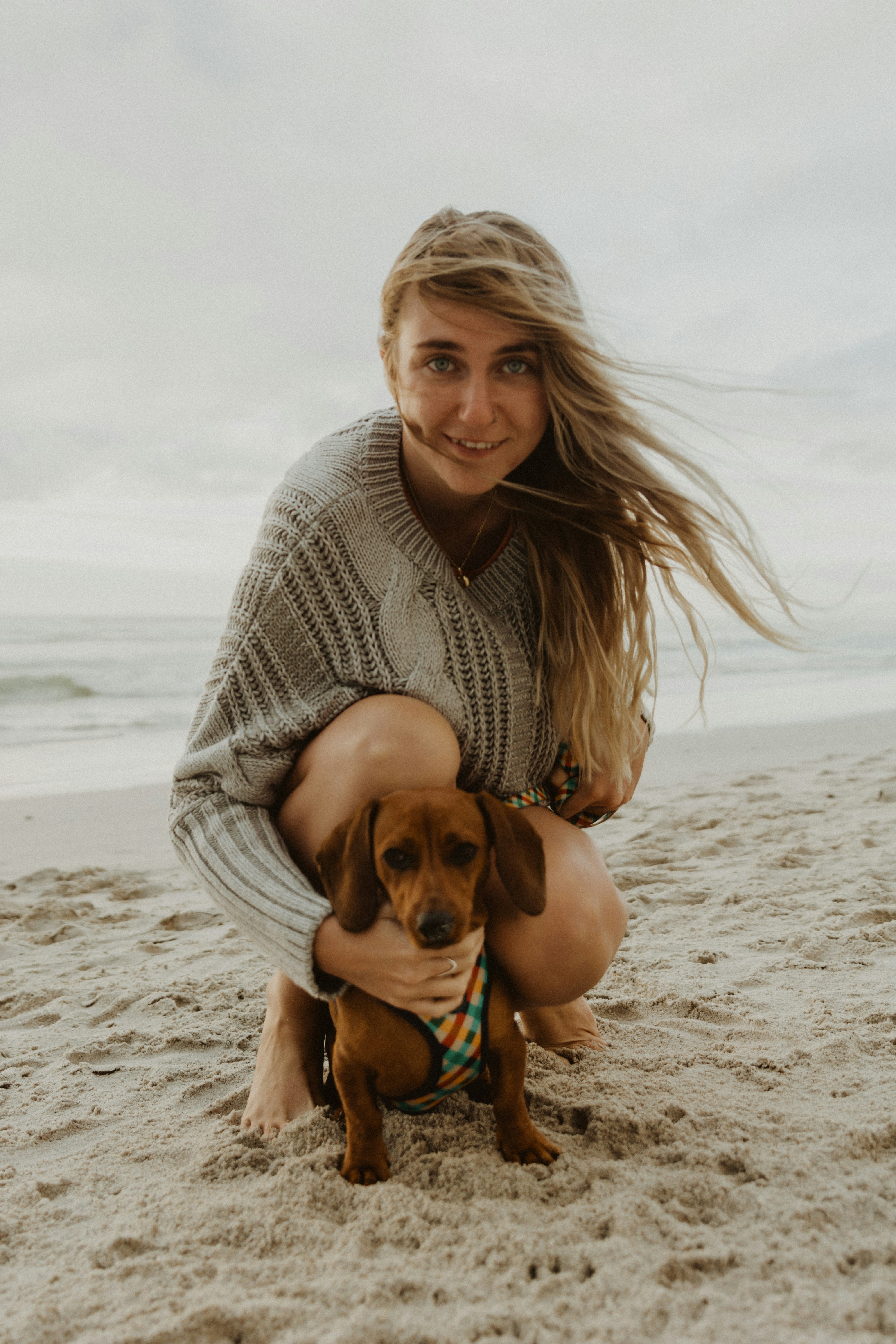 woman in gray knit sweater hugging brown short coated dog on beach during daytime