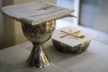 A decorative silver chalice with intricate engravings sits on a white tablecloth. A folded white cloth with embroidered designs is placed on top of a similar ornate silver bowl. Next to it is a card with a cross design in red and gold.