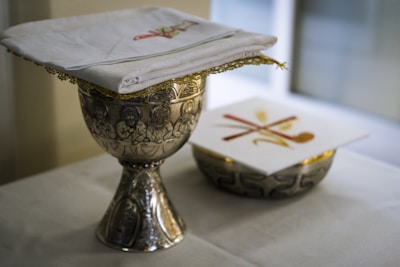 A decorative silver chalice with intricate engravings sits on a white tablecloth. A folded white cloth with embroidered designs is placed on top of a similar ornate silver bowl. Next to it is a card with a cross design in red and gold.