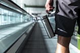 A person holding a stainless steel water bottle is walking on an airport moving walkway. The focus is on the lower half of the body, wearing shorts. The background is blurred, emphasizing the glass walls and the long walkway.