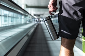 A person holding a stainless steel water bottle is walking on an airport moving walkway. The focus is on the lower half of the body, wearing shorts. The background is blurred, emphasizing the glass walls and the long walkway.