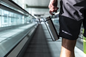 A person holding a stainless steel water bottle is walking on an airport moving walkway. The focus is on the lower half of the body, wearing shorts. The background is blurred, emphasizing the glass walls and the long walkway.