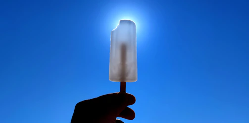 Hand holding a bright red strawberry popsicle against a clear blue sky