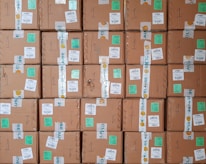 A stack of cardboard boxes with various labels and stickers, arranged in a neat and organized manner. Each box has handwritten numbers and multiple shipping labels, some of which are on tape that seals the boxes.