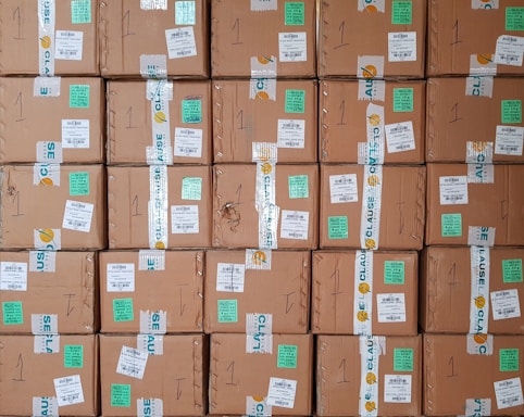 A stack of cardboard boxes with various labels and stickers, arranged in a neat and organized manner. Each box has handwritten numbers and multiple shipping labels, some of which are on tape that seals the boxes.