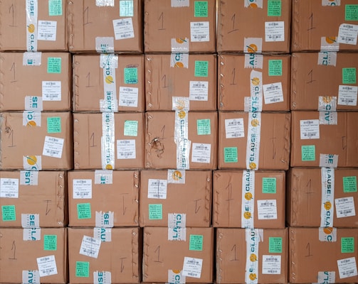 A stack of cardboard boxes with various labels and stickers, arranged in a neat and organized manner. Each box has handwritten numbers and multiple shipping labels, some of which are on tape that seals the boxes.