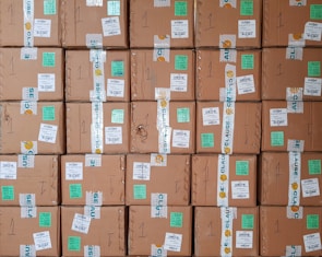 A stack of cardboard boxes with various labels and stickers, arranged in a neat and organized manner. Each box has handwritten numbers and multiple shipping labels, some of which are on tape that seals the boxes.