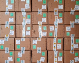 A stack of cardboard boxes with various labels and stickers, arranged in a neat and organized manner. Each box has handwritten numbers and multiple shipping labels, some of which are on tape that seals the boxes.