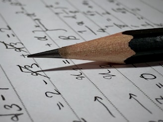 A sharpened pencil lying on a sheet of paper filled with handwritten mathematical equations and symbols. The paper appears to be lined with faint horizontal dots, and the pencil is positioned diagonally across the page, with its point directed towards some of the written equations.