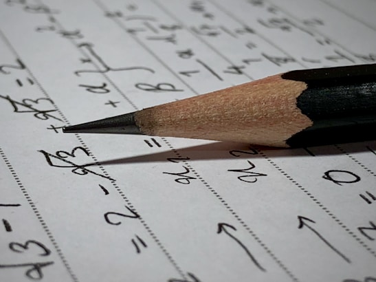 A sharpened pencil lying on a sheet of paper filled with handwritten mathematical equations and symbols. The paper appears to be lined with faint horizontal dots, and the pencil is positioned diagonally across the page, with its point directed towards some of the written equations.
