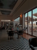 An indoor dining area with a modern and stylish design, featuring zigzag patterned flooring, dark chairs and tables, and large windows that let in natural light. A person is standing near a counter, and there are various decorative elements visible.