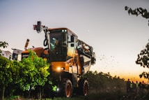 A large agricultural machine navigates through a vineyard at dusk. The machine is orange with black details, and bright lights illuminate the surrounding grapevines. Several people stand nearby, watching the operation as the sky transitions from daytime to evening.