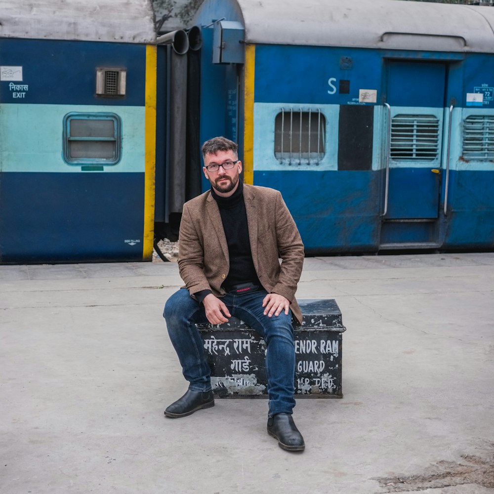 昼間、青い電車のそばの黒い金属製のベンチに座る黒いブレザーの男