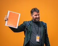 A man with a beard, wearing a dark blazer and turtleneck, holds up an orange booklet labeled 'freewalking Tour Salzburg' against a bright orange background. He is smiling and carrying a backpack with a visible name tag that matches the booklet's color scheme.