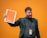 A man with a beard, wearing a dark blazer and turtleneck, holds up an orange booklet labeled 'freewalking Tour Salzburg' against a bright orange background. He is smiling and carrying a backpack with a visible name tag that matches the booklet's color scheme.