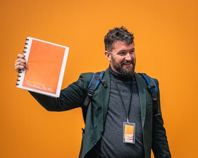 A man with a beard, wearing a dark blazer and turtleneck, holds up an orange booklet labeled 'freewalking Tour Salzburg' against a bright orange background. He is smiling and carrying a backpack with a visible name tag that matches the booklet's color scheme.