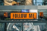 A bright orange sign with the words 'FOLLOW ME' is mounted on top of a vehicle. The sign is flanked by two orange warning lights. In the background, the glass facade of a building reflects the surroundings, adding an urban atmosphere.