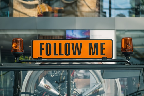 A bright orange sign with the words 'FOLLOW ME' is mounted on top of a vehicle. The sign is flanked by two orange warning lights. In the background, the glass facade of a building reflects the surroundings, adding an urban atmosphere.