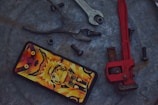 A smartphone with a vibrant anime-themed case featuring a character in bold orange and yellow hues is placed on a rough metallic surface. Surrounding the phone are various tools and hardware, including a set of pliers, an open-ended wrench, a red pipe wrench, and several black bolts and a nut.