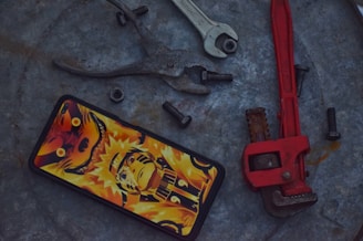 A smartphone with a vibrant anime-themed case featuring a character in bold orange and yellow hues is placed on a rough metallic surface. Surrounding the phone are various tools and hardware, including a set of pliers, an open-ended wrench, a red pipe wrench, and several black bolts and a nut.