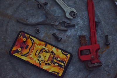 A smartphone with a vibrant anime-themed case featuring a character in bold orange and yellow hues is placed on a rough metallic surface. Surrounding the phone are various tools and hardware, including a set of pliers, an open-ended wrench, a red pipe wrench, and several black bolts and a nut.