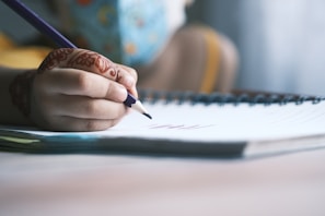 A hand with intricate henna designs is holding a purple pencil, writing on a lined notebook. The focus is on the hand and pencil, while the background is softly blurred, suggesting an indoor setting. The notebook lies on a table, and part of a person's clothing is slightly visible in the background.