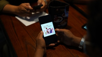 A person is holding a smartphone displaying a graphic with text, while another person in the background is using a phone. The setting appears to be a table with a menu or advertisement stand.