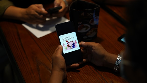 A person is holding a smartphone displaying a graphic with text, while another person in the background is using a phone. The setting appears to be a table with a menu or advertisement stand.
