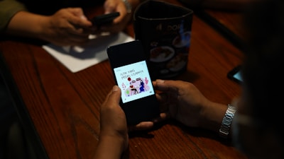 A person is holding a smartphone displaying a graphic with text, while another person in the background is using a phone. The setting appears to be a table with a menu or advertisement stand.