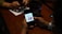 A person is holding a smartphone displaying a graphic with text, while another person in the background is using a phone. The setting appears to be a table with a menu or advertisement stand.
