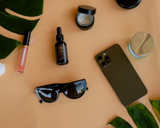 A flat lay composition featuring various objects on a light brown surface. Items include sunglasses, a smartphone, a bottle of oil or serum, an open jar of cream, a closed jar with a silver lid, a lip gloss, and large green leaves. The arrangement suggests a theme of lifestyle or beauty products.