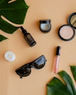 Various beauty and lifestyle items arranged aesthetically on a beige background. The items include a glass bottle with a dropper, an open jar of cream, a compact powder, a white candle, black sunglasses, a lip gloss, and large green leaves placed diagonally.