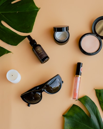 Various beauty and lifestyle items arranged aesthetically on a beige background. The items include a glass bottle with a dropper, an open jar of cream, a compact powder, a white candle, black sunglasses, a lip gloss, and large green leaves placed diagonally.